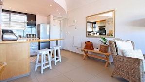 A modern kitchen with white cabinets, a stainless steel refrigerator, and a wooden countertop. There are two white stools and a wicker sofa with cushions. A mirror hangs on the wall, reflecting the kitchen. - Morakani (Cape Town)