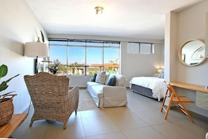 A modern bedroom with a bed, a sofa, a wicker chair, a desk, and a large window offering a view of buildings and trees.