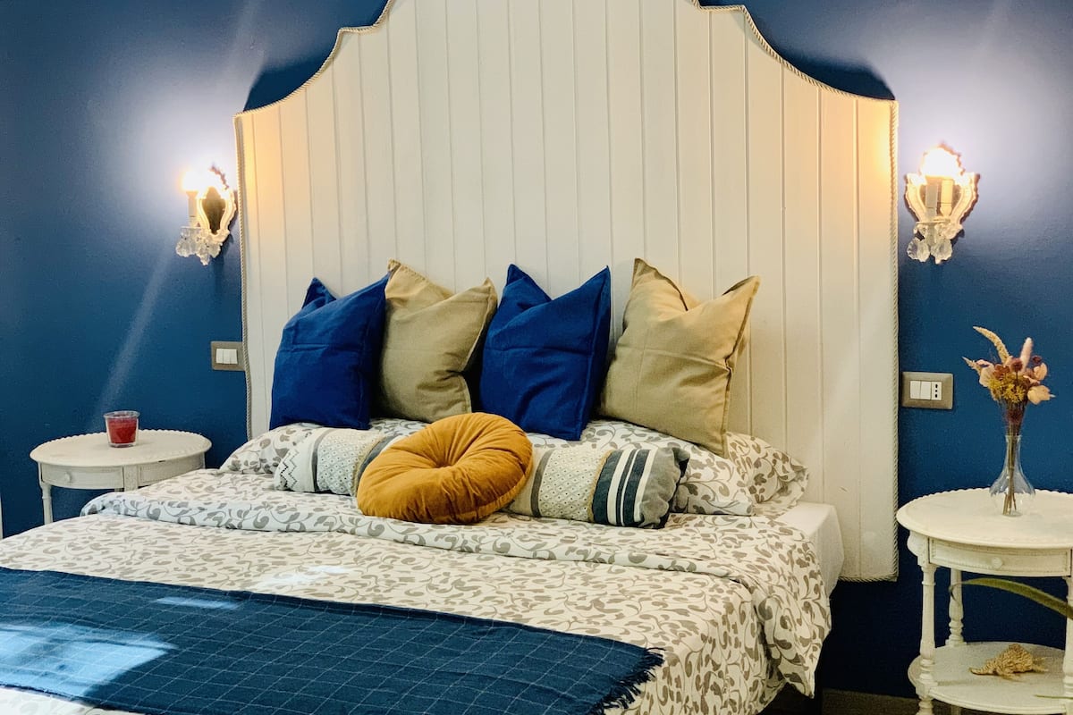 Ein Schlafzimmer mit Bett, Kissen, einem Nachttisch, einer Lampe und einer Vase mit Blumen.