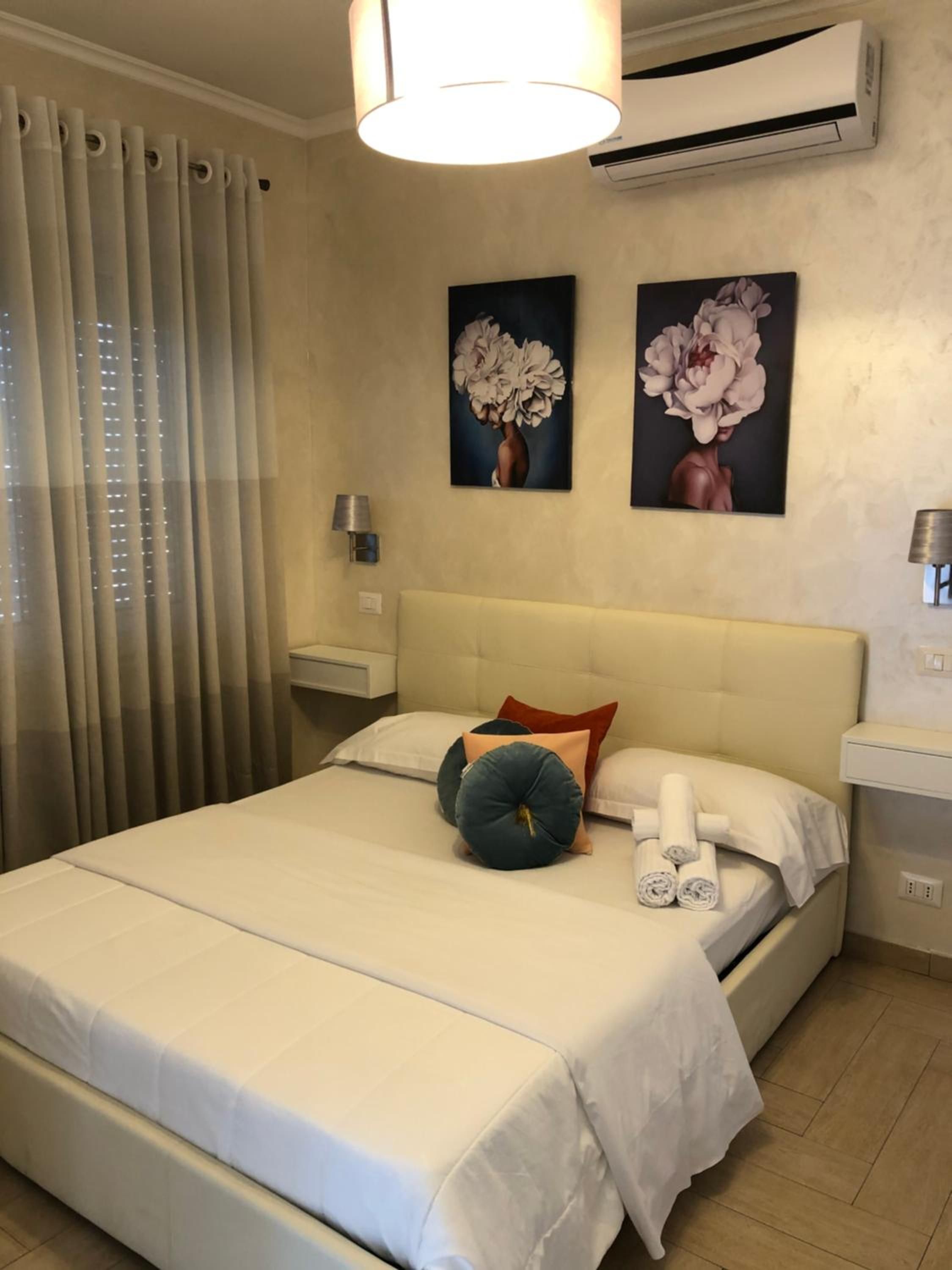A hotel room with a bed, curtains, an air conditioning unit, a wall-mounted lamp, and two paintings.