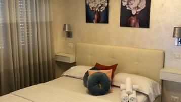 A hotel room with a bed, curtains, an air conditioning unit, a wall-mounted lamp, and two paintings.