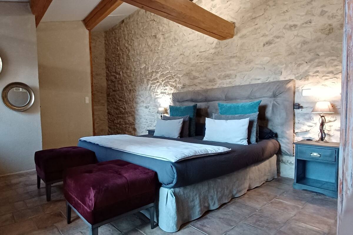 Een slaapkamer met een stenen muur, een bed met veel kussens, een nachtkastje met een lamp en een klein bankje.