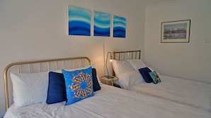 Two single beds with white bedding and blue pillows, a bedside table with a lamp, and wall art above the beds. - Birch at Somerhill (Hove)