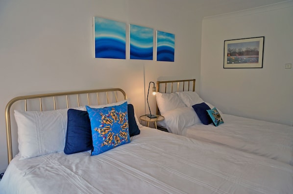 Two single beds with white bedding and blue pillows, a bedside table with a lamp, and wall art above the beds.