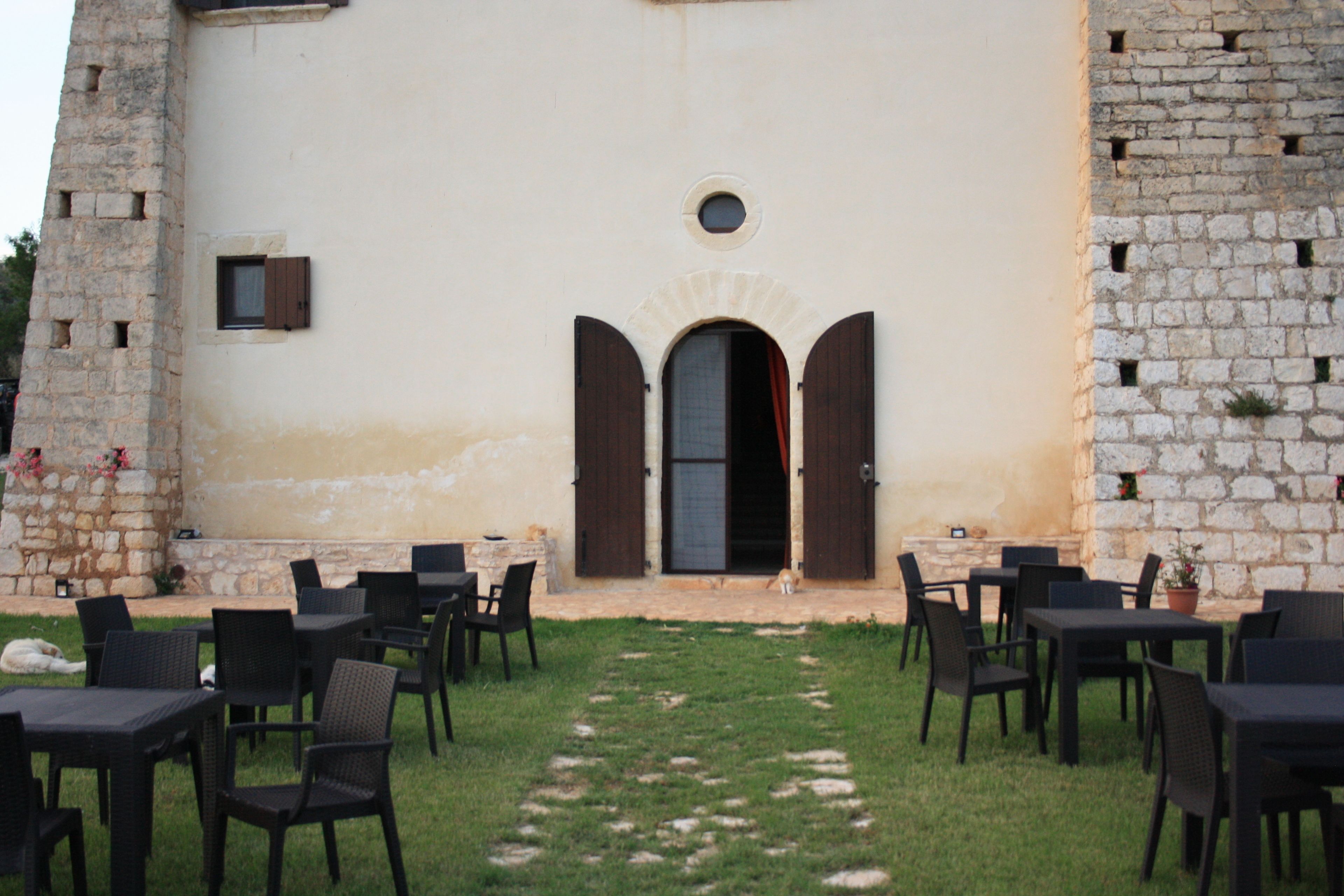 Un espace extérieur aménagé avec des tables et des chaises, situé devant un bâtiment à la façade en pierre et aux entrées en arc de cercle.
