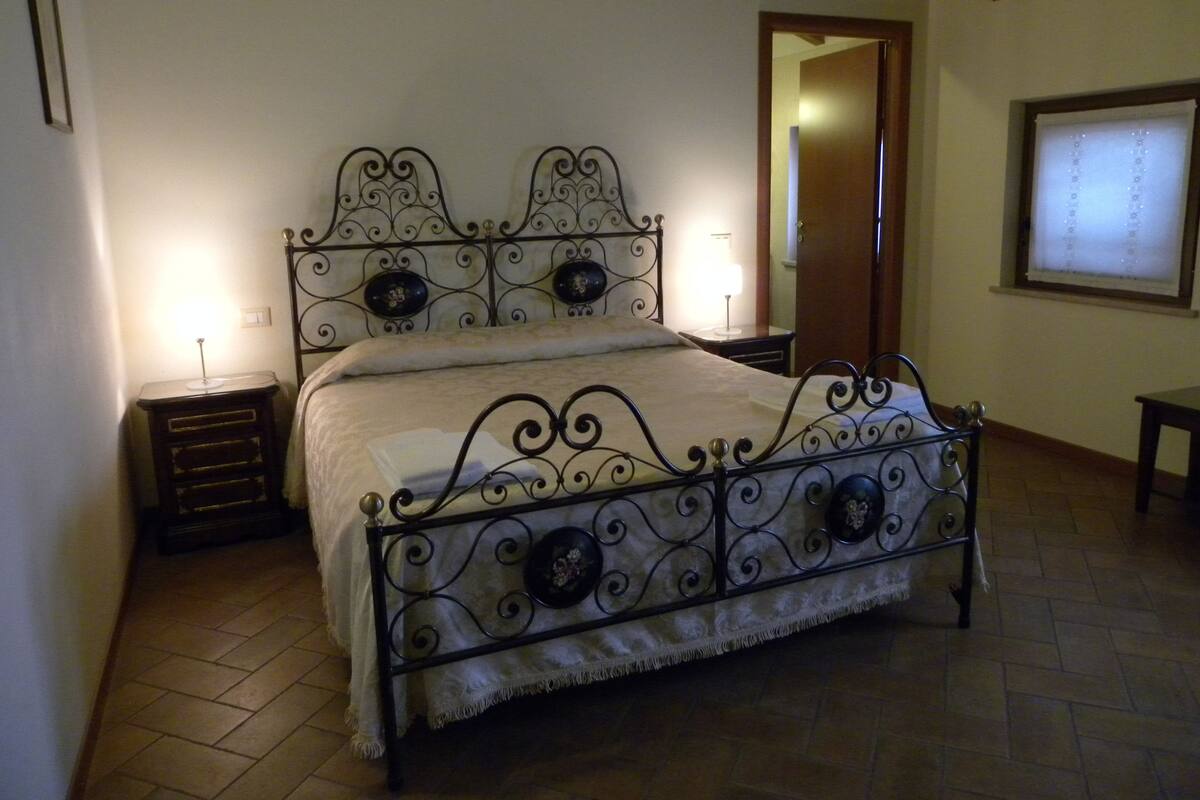Un dormitorio con una cama de estructura metálica, dos mesitas de noche, un velador y una ventana con persianas, además de una puerta que da a otra habitación.