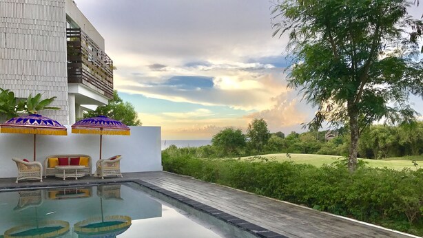A pool area with a view of the sea, surrounded by greenery and a white building.