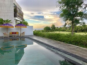 Área de piscina com vista para o mar, cercada por vegetação e um edifício branco.