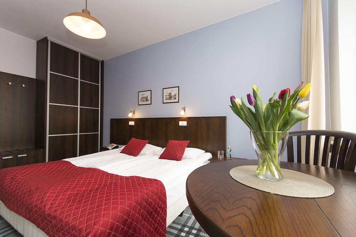 Habitación de hotel con una cama grande, una mesita de noche de madera, un florero con flores y una mesa de comedor con un mantel individual.