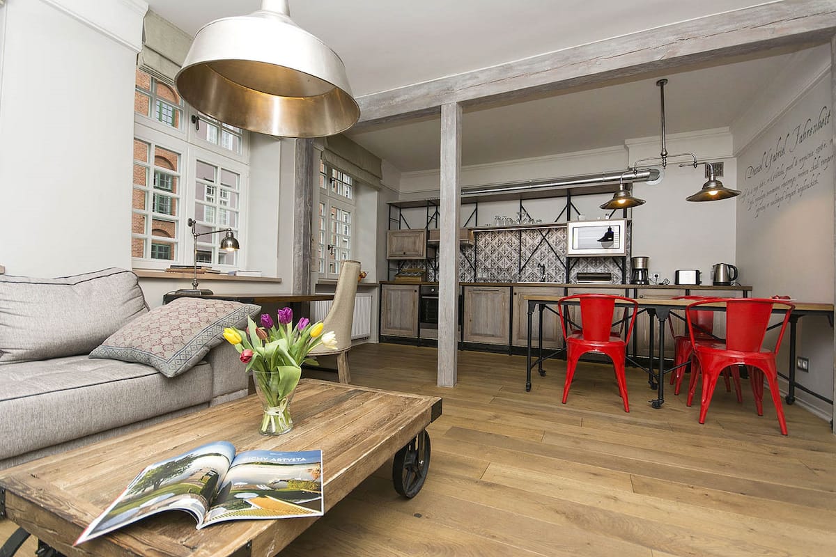 Amplia sala de estar con un sofá, una mesa de centro con revistas, un jarrón de flores y una cocina al fondo.