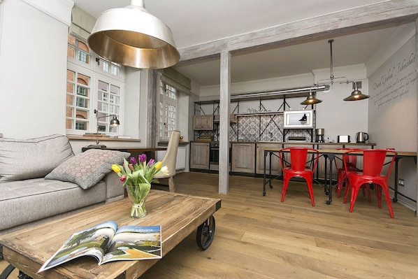 A spacious living area with a sofa, a coffee table with magazines, a vase of flowers, and a kitchen in the background.