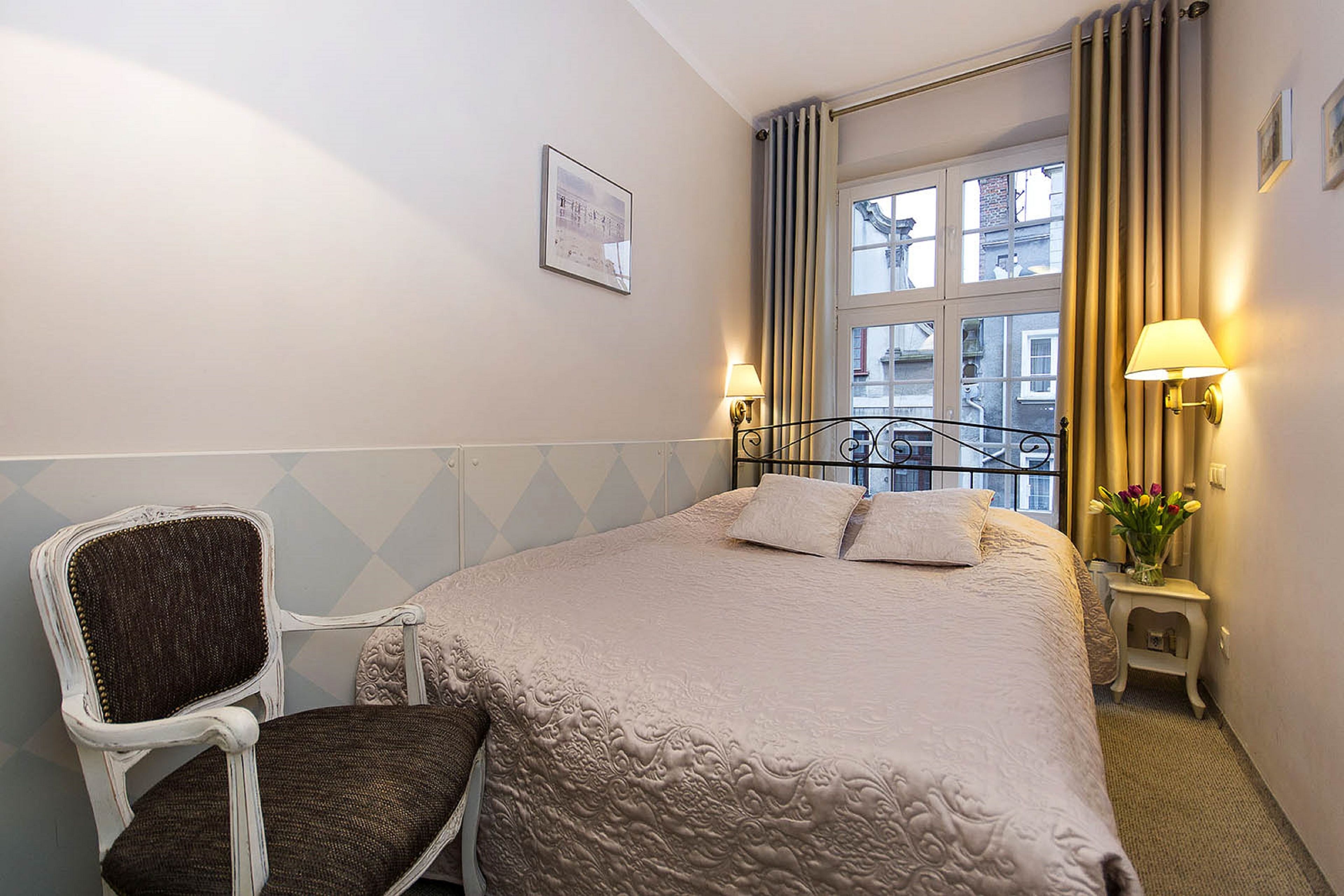 A hotel room with a bed, a chair, a window with curtains, and a small table with a lamp and flowers.
