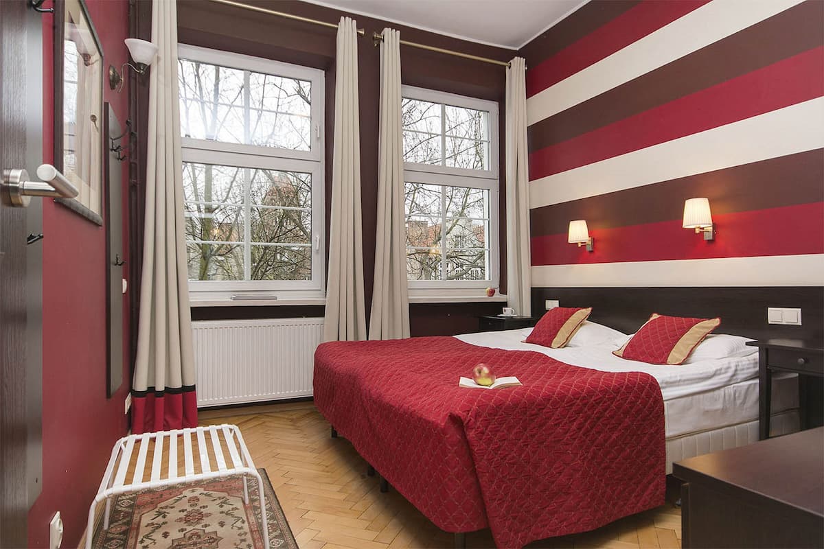 Una habitación de hotel con una cama grande, dos ventanas con cortinas, una mesita de noche y un buró.