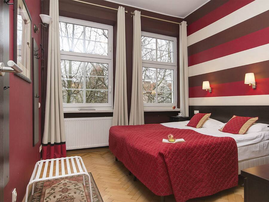 Una habitación de hotel con una cama grande, dos ventanas con cortinas, una mesita de noche y un buró.