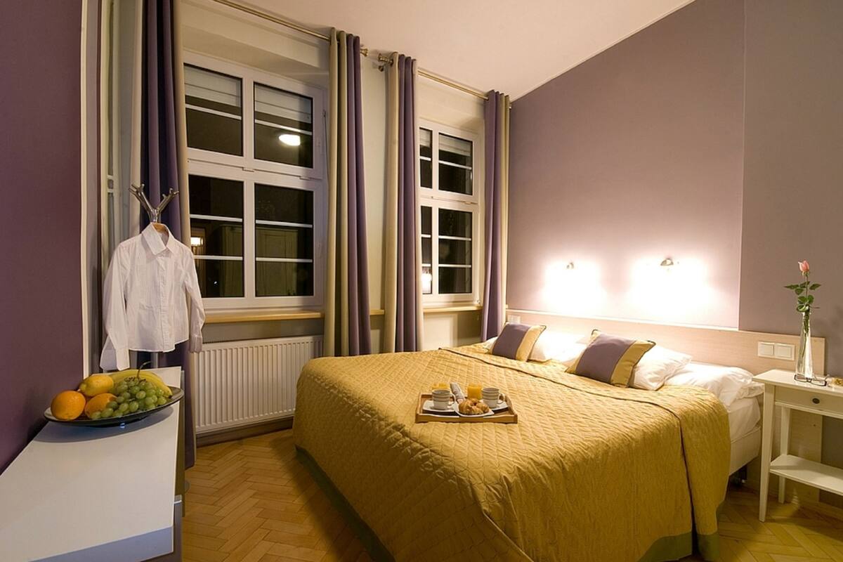 Habitación de hotel con cama, mesita de noche y ventana con cortinas. Hay una bandeja con productos de desayuno sobre la cama.