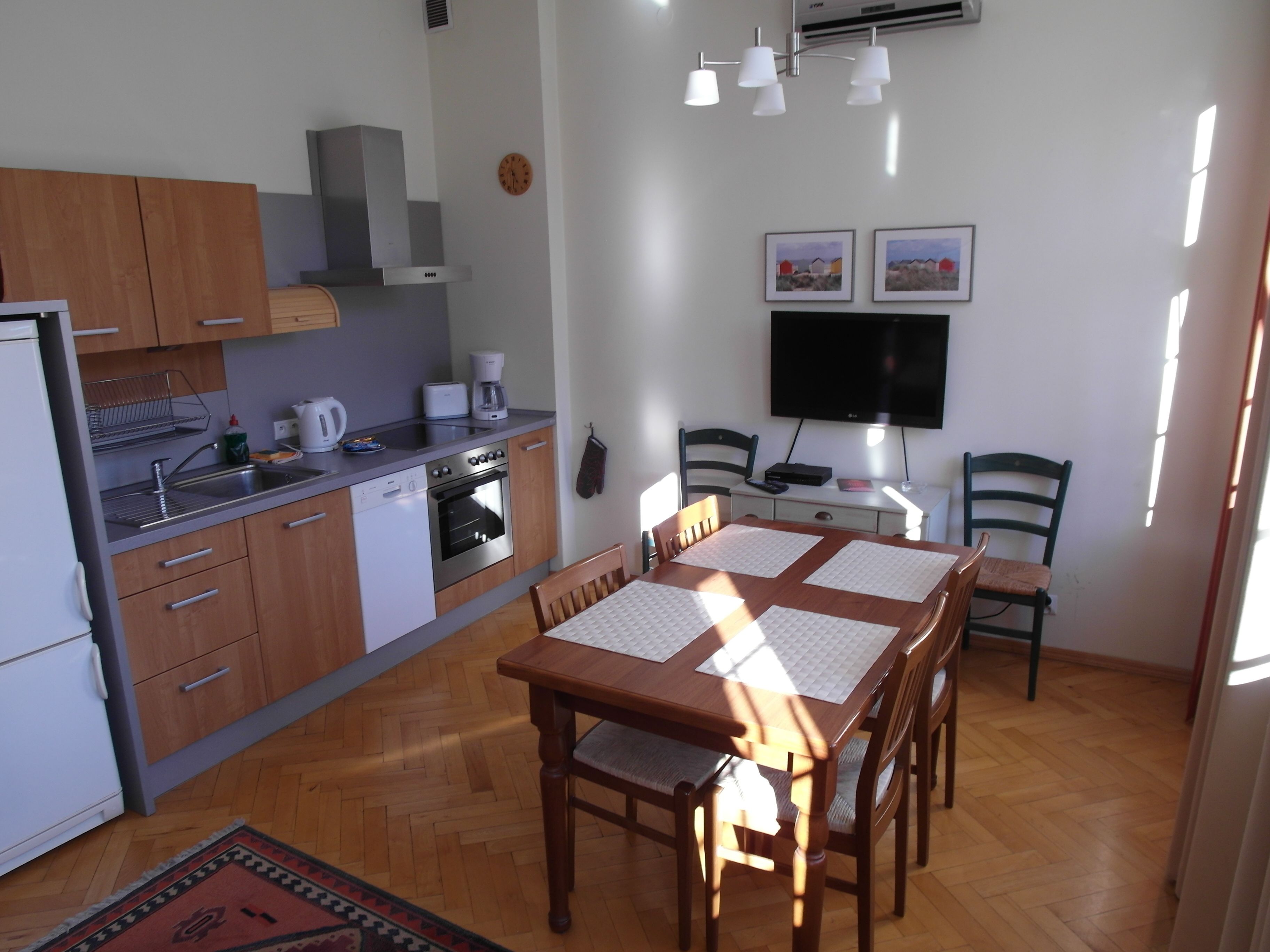 Cocina compacta con lavavajillas, estufa y cafetera. Hay una mesa de comedor con cuatro sillas y un televisor montado en la pared.