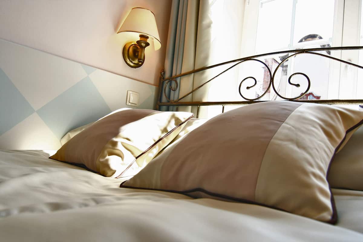 Una cama bien hecha con cabecera, una lámpara de pared y una ventana con cortinas.