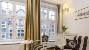 A room with a large window, a white radiator, a striped sofa, a white cabinet, and a small table with a vase of flowers. - Patio Apartamenty (Gdansk)