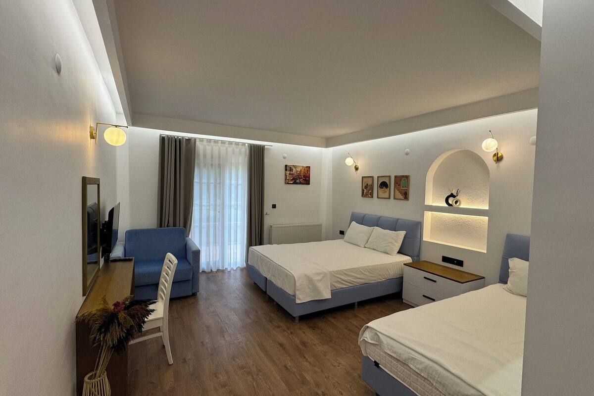 Habitación de hotel con dos camas, un sofá, un escritorio con una silla y un ventanal con cortinas.