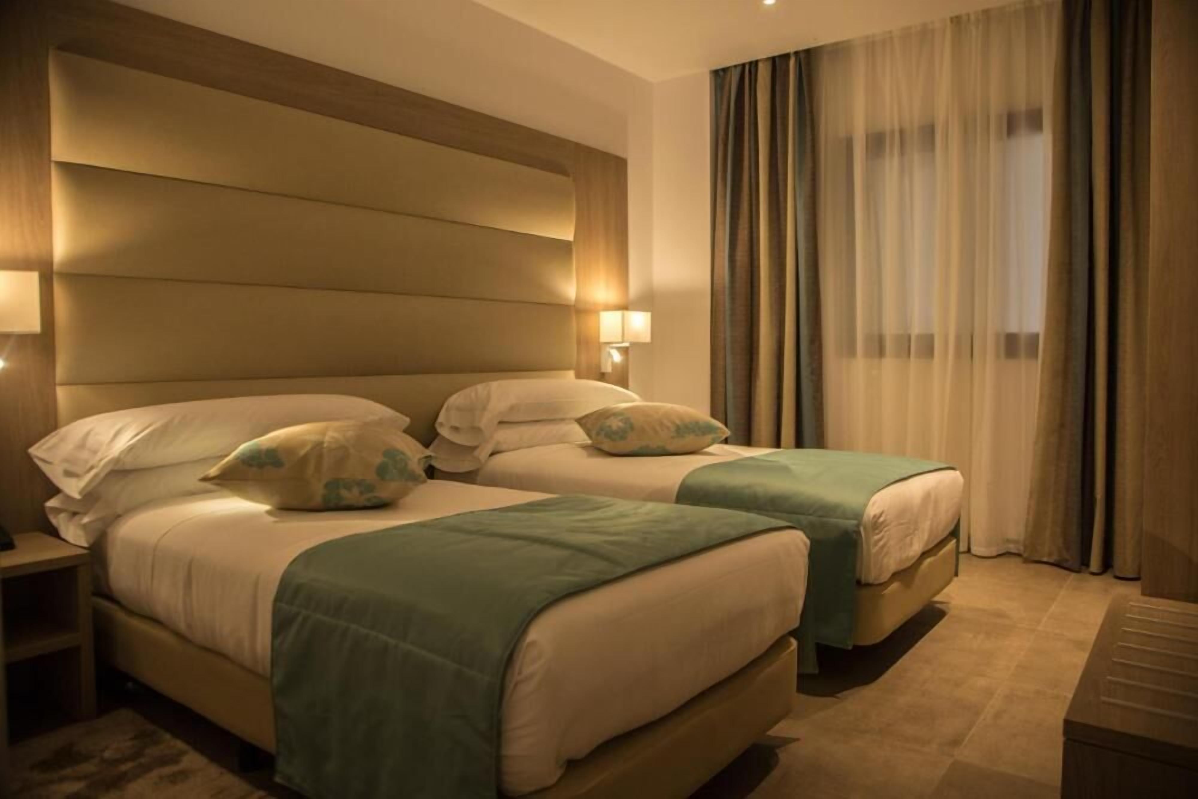 Una habitación de hotel con dos camas, un cabecero grande, una mesita de noche y una ventana con cortinas.