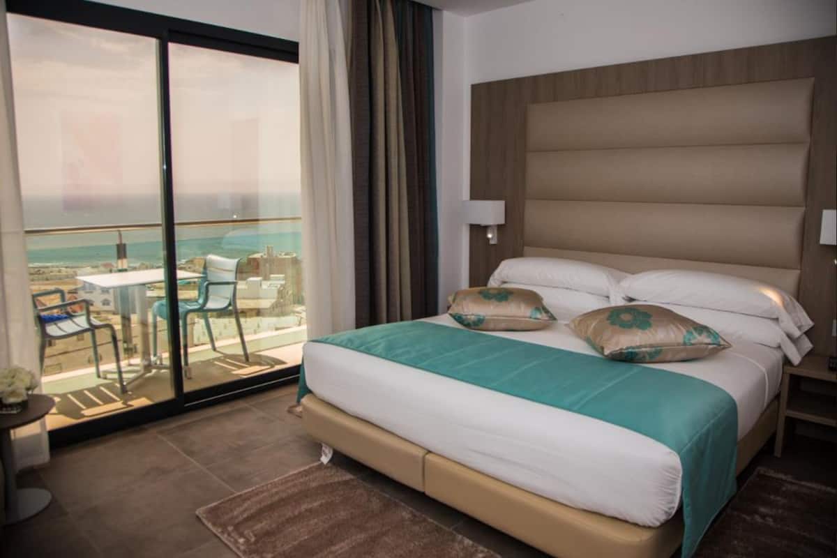 Een hotelkamer met een groot bed, een balkon met uitzicht op zee en een kleine tafel met stoelen.