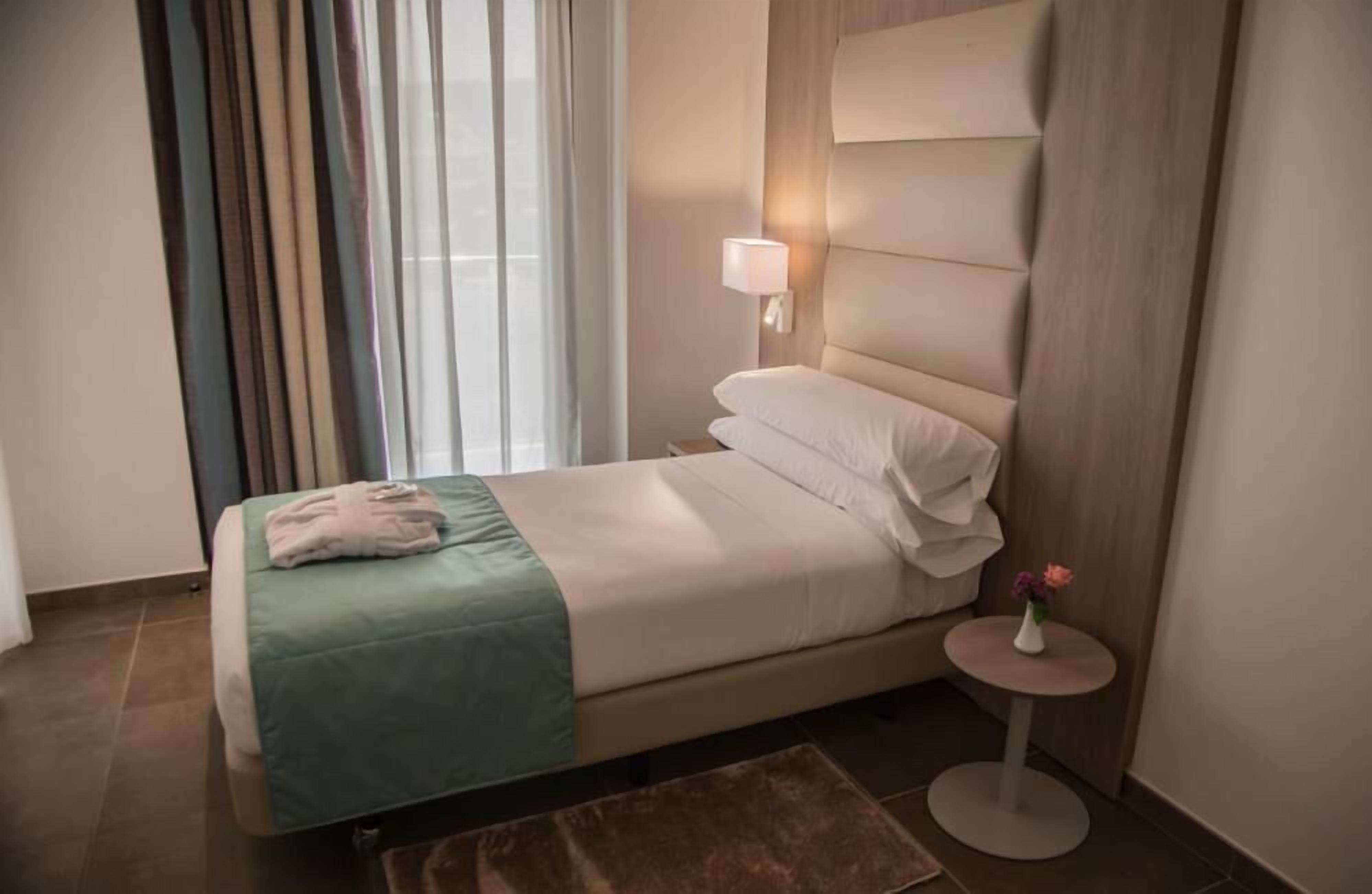 Une chambre d’hôtel avec un lit, une table de chevet avec une lampe, une fenêtre avec des rideaux et une petite table sur laquelle est posé un vase.