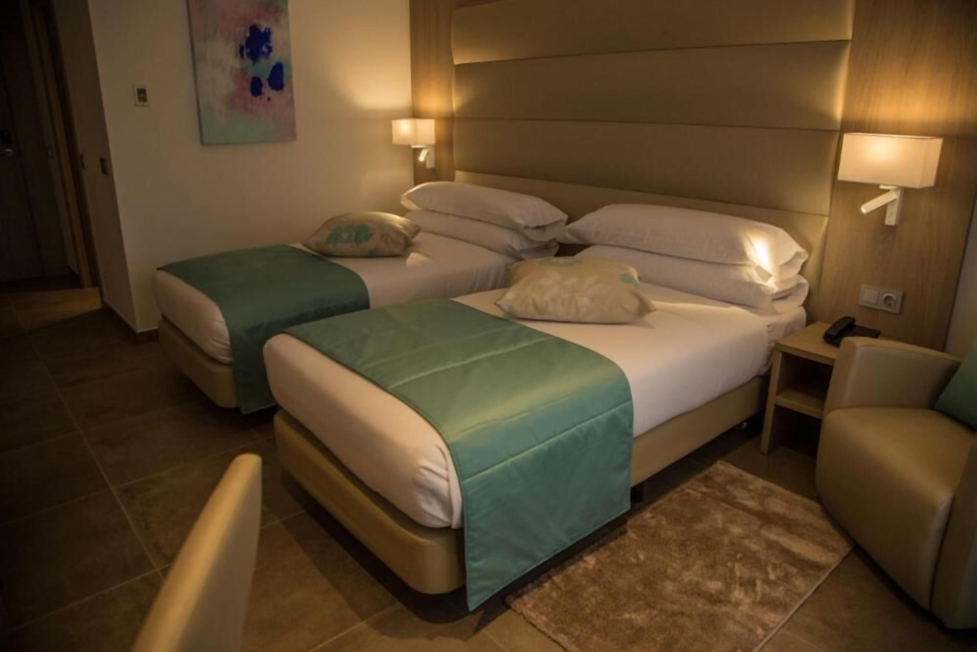 Habitación de hotel con una cama grande, dos camas más pequeñas, un sofá y una mesita de noche con lámpara.