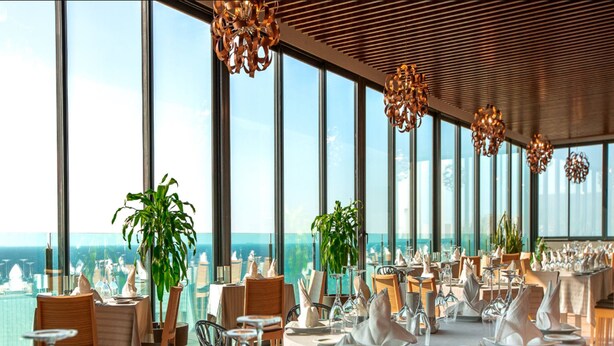 A restaurant with tables set for dining, large windows offering a view of the ocean, and hanging pendant lights. - Az Hotel Le Zephyr Mostaganem (Mazagran)