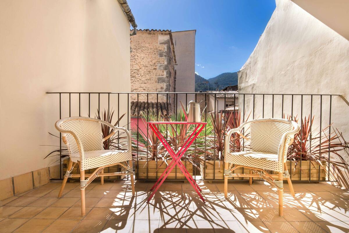 A balcony with two wicker chairs, a red metal table, and potted plants.