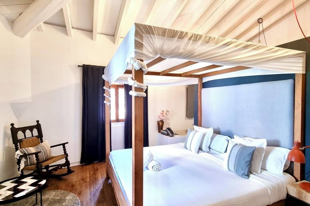 A four-poster bed with a canopy, a rocking chair, a bedside table with a lamp, and a wooden floor.