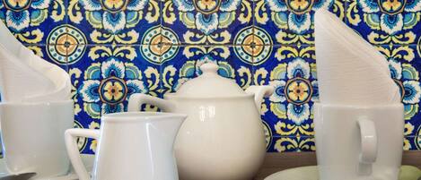 A table set with white ceramic tea set including a teapot, cups, and a sugar bowl, against a blue and yellow patterned tile wall.