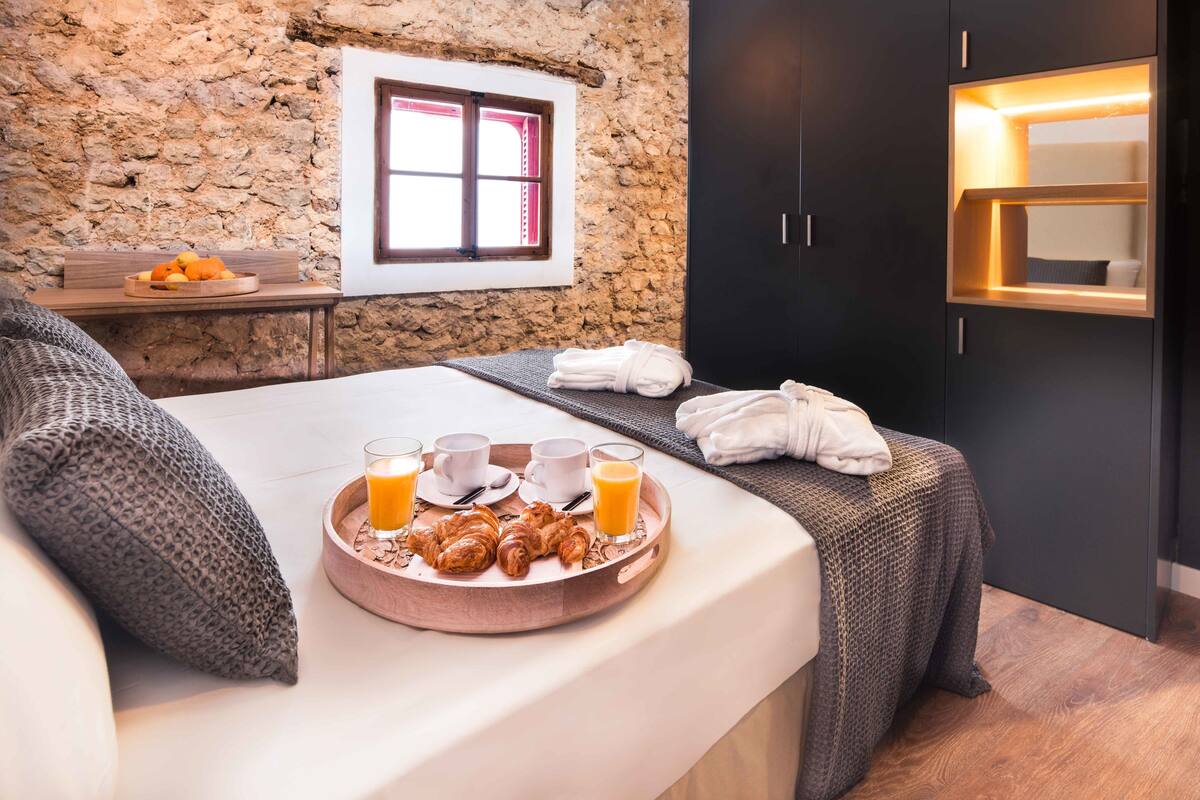A bedroom with a stone wall, a window, a bedside table with breakfast items, and a bed with a blanket.