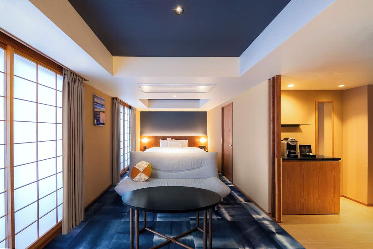 A hotel room with a bed, a small table, a kitchenette, and a view of the outside through large windows.
