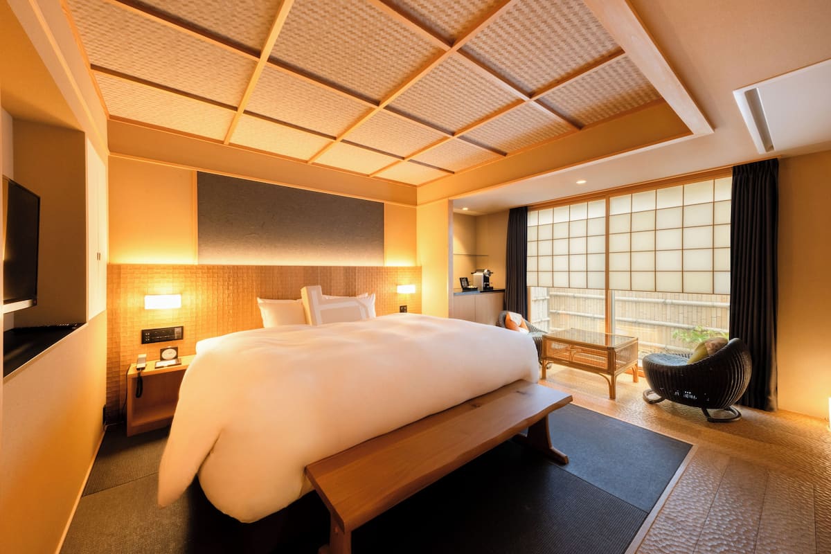 A hotel room with a large bed, a bench, a TV, a bedside table, a lamp, and a window with a view.
