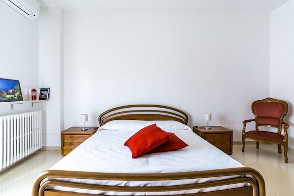 Un dormitorio ordenado con una cama de madera, un cojín rojo, una mesita de noche de madera con una lámpara, una silla de madera y un televisor montado en la pared.