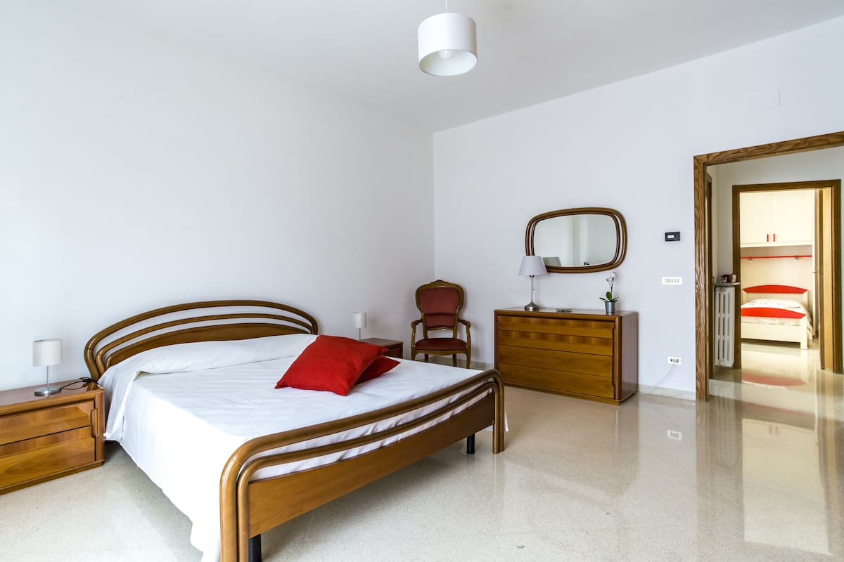 Quarto com cama de madeira, mesa de cabeceira, cômoda, cadeira e espelho na parede.