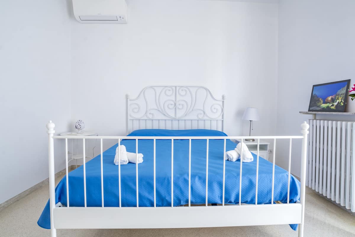 Uma cama de metal branca com colchão azul e almofadas brancas, uma mesinha de cabeceira com abajur, e um porta-retrato na parede.