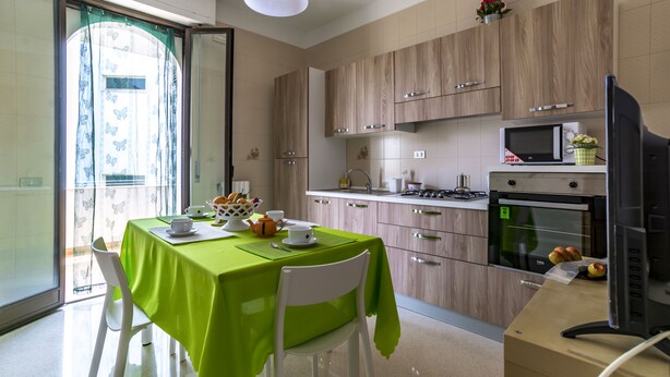 A modern kitchen with wooden cabinets, a dining table with a green tablecloth, and a TV on a stand. - Appartamenti Albero di Giada (Polignano a Mare)