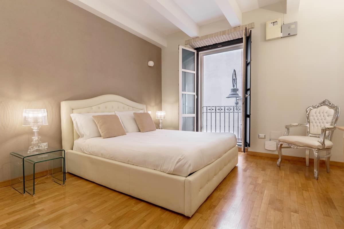 A bedroom with a bed, bedside table, lamp, chair, and an open door leading to a balcony.