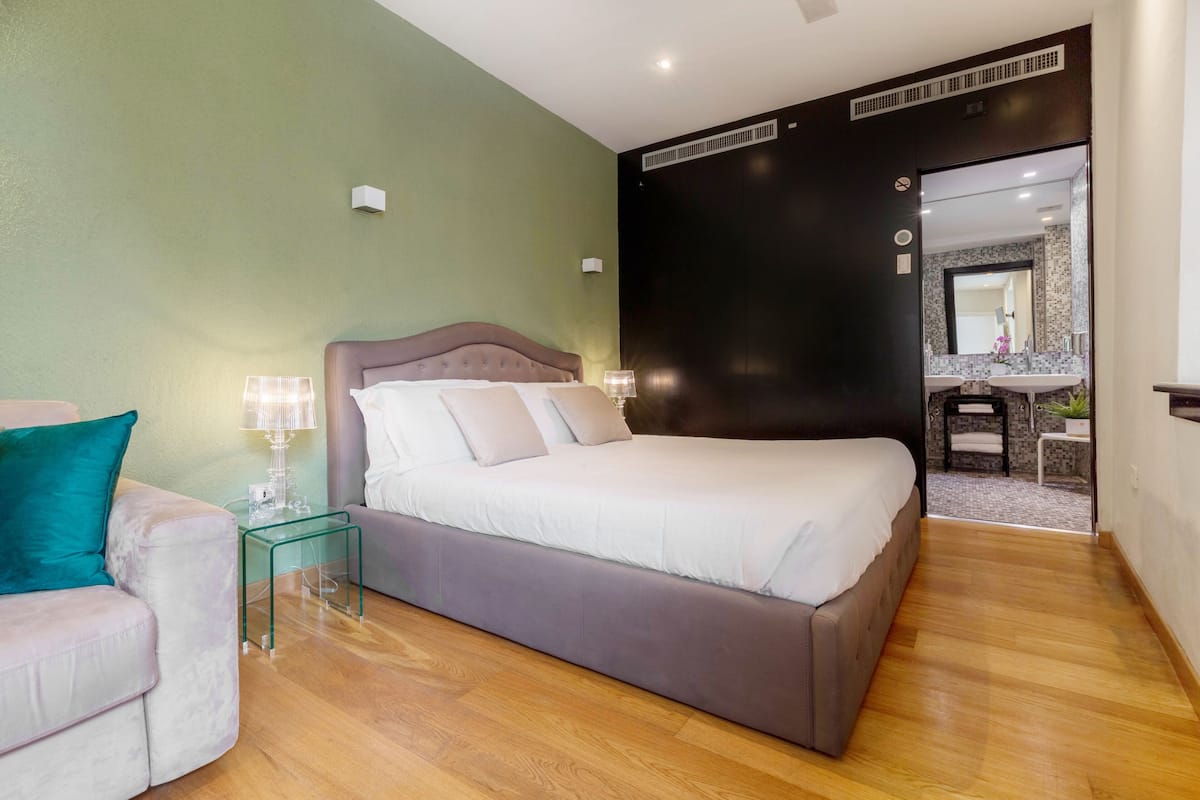 A modern bedroom with a large bed, a glass side table, a chair, and a bathroom visible through an open door.