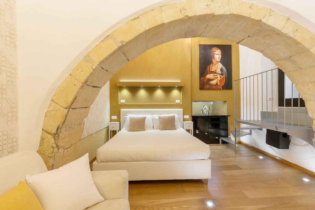 A bedroom with a large bed, a sofa, an arched stone ceiling, and a painting on the wall.