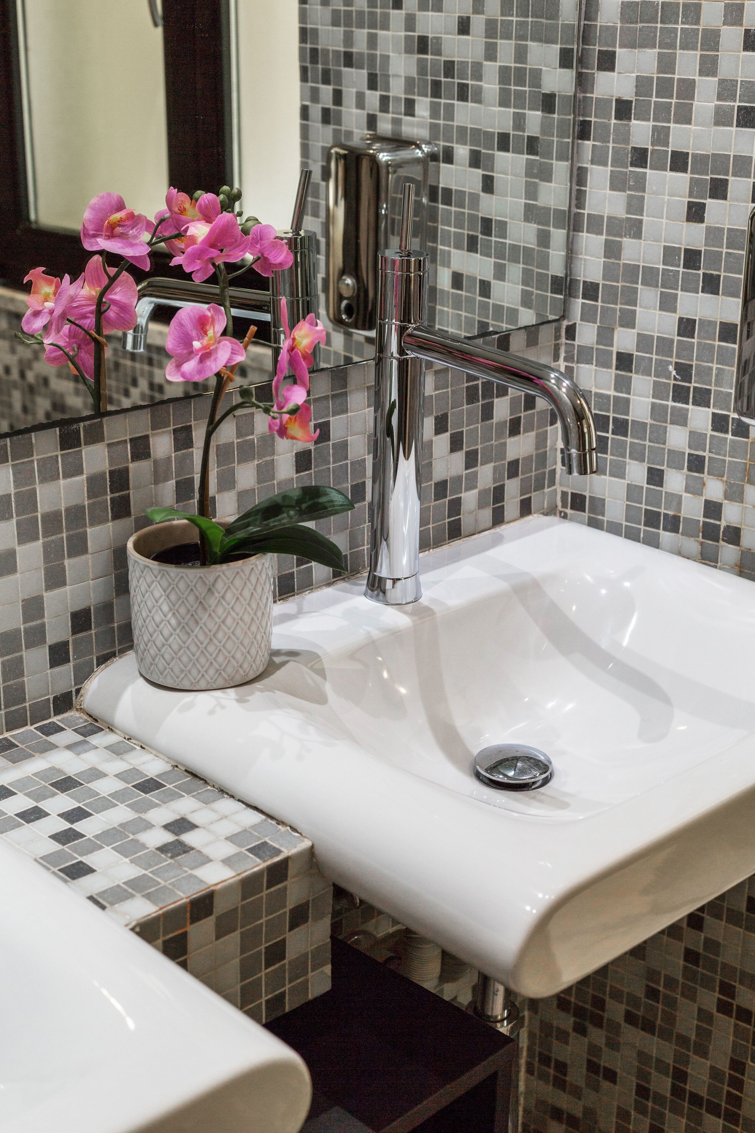 Ein Badezimmer mit einem weißen Waschbecken, einem modernen Wasserhahn und einer Orchidee im Topf. Die Wände sind mit einem Mosaikmuster gefliest.