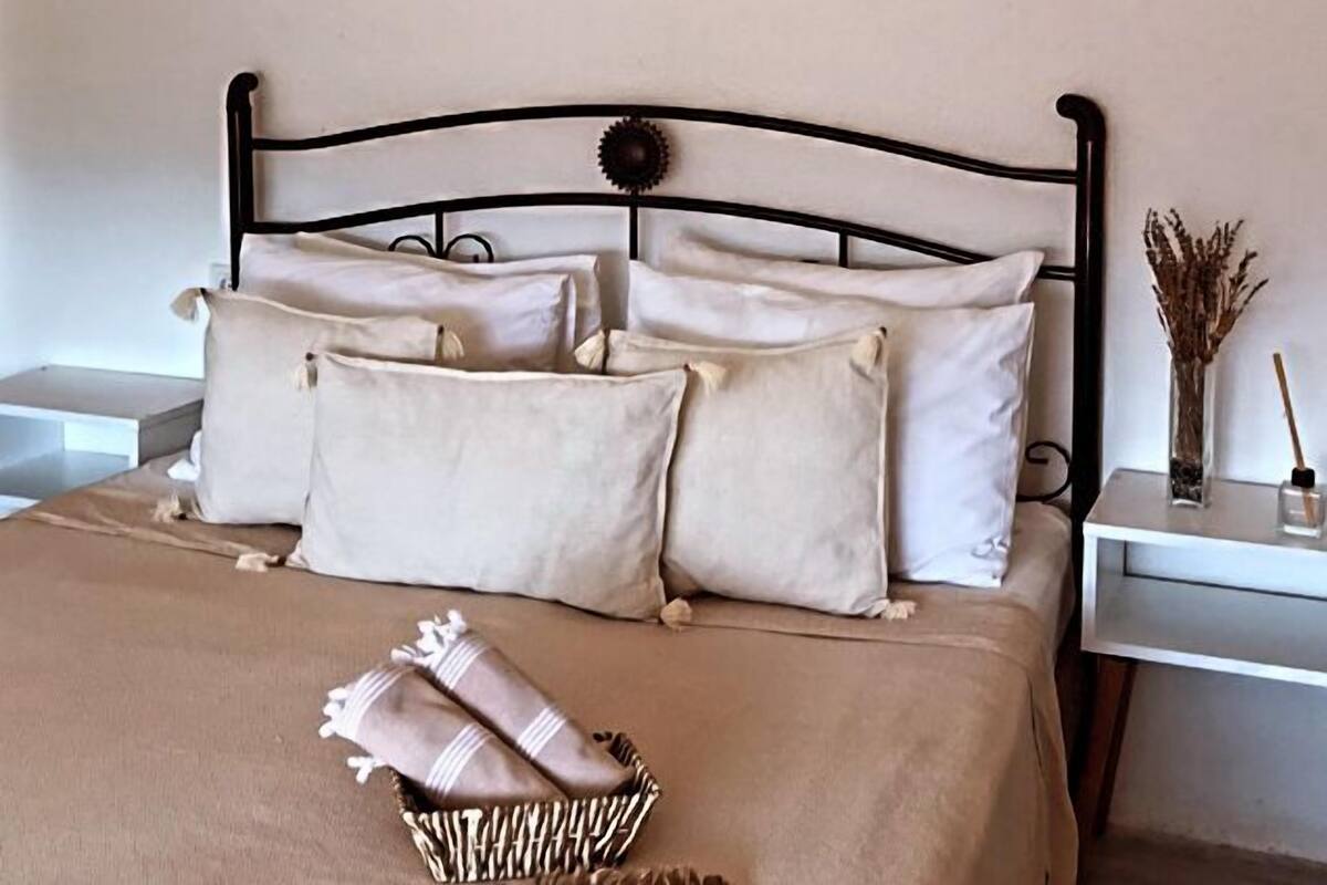 A neatly made bed with a beige bedspread and pillows, a basket with folded towels, and a wall-mounted picture of a bicycle.