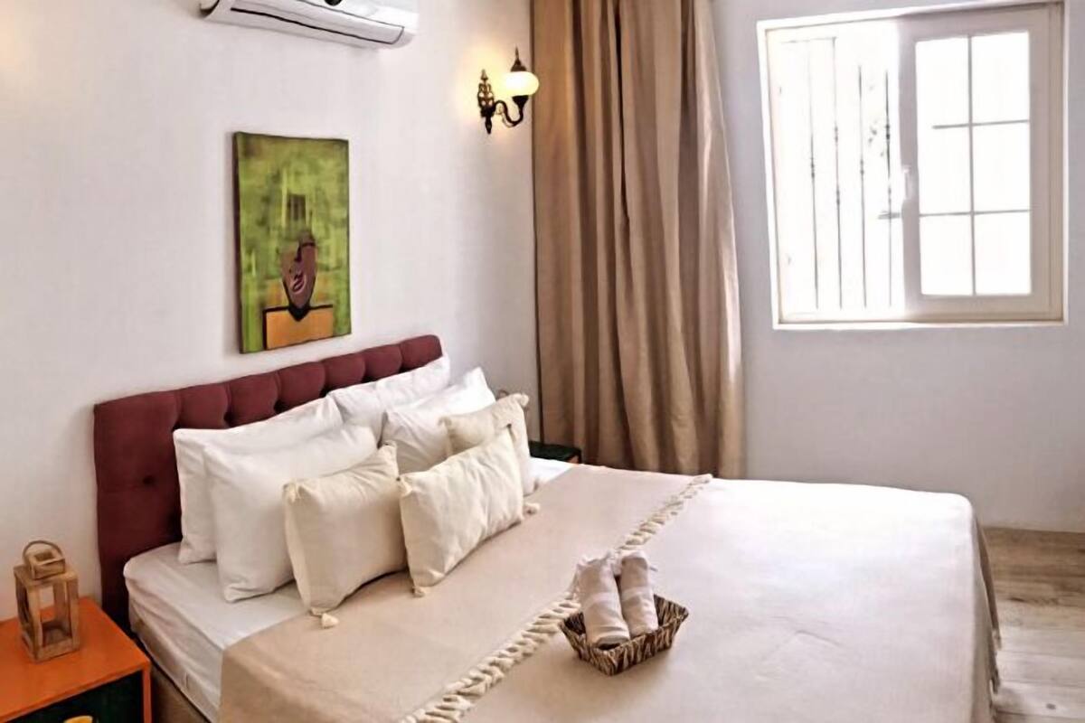 A neatly made bed with white linens and a red headboard, a bedside lamp, and a painting on the wall.