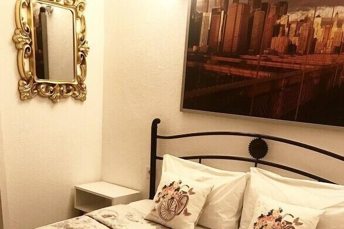 A neatly made bed with floral bedding and two decorative pillows, a bedside table, a wall-mounted mirror, and a large framed picture of a cityscape.
