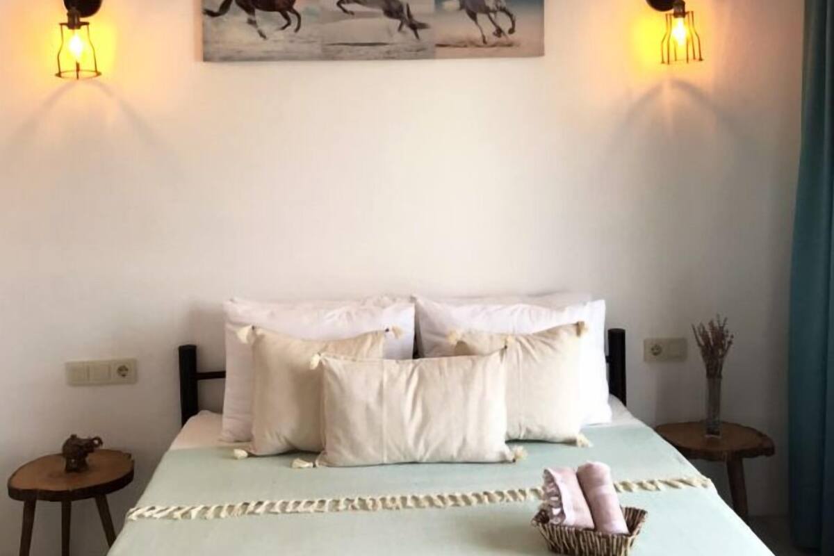 A bedroom with a bed, pillows, a bedside table, a wall-mounted painting, and wall sconces.