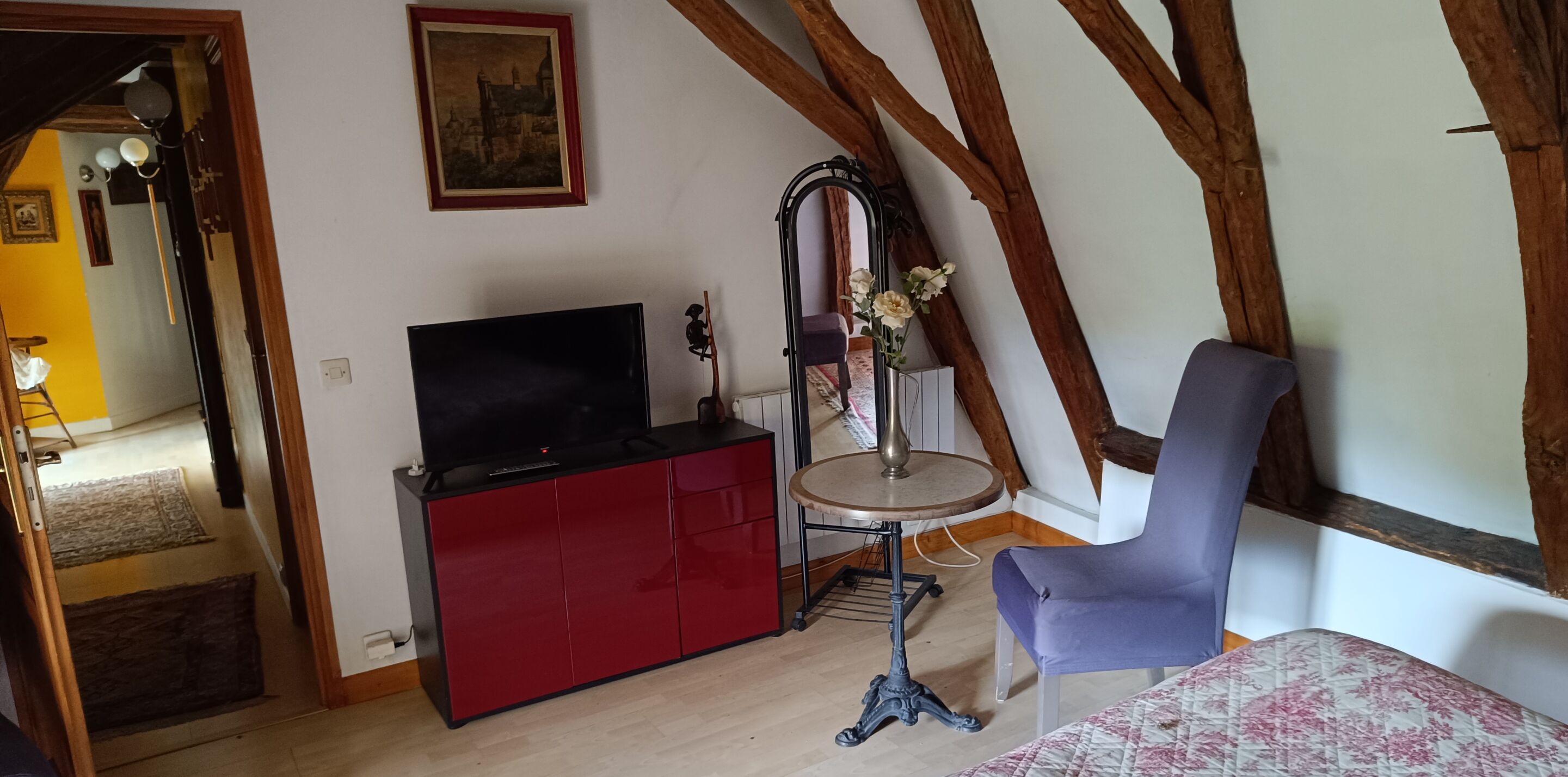 Ein Zimmer mit einer roten Unterhaltungsecke, einem Fernseher, einem kleinen runden Tisch mit Blumen in einer Vase, einem blauen Sessel und sichtbaren Holzbalken an der Decke.
