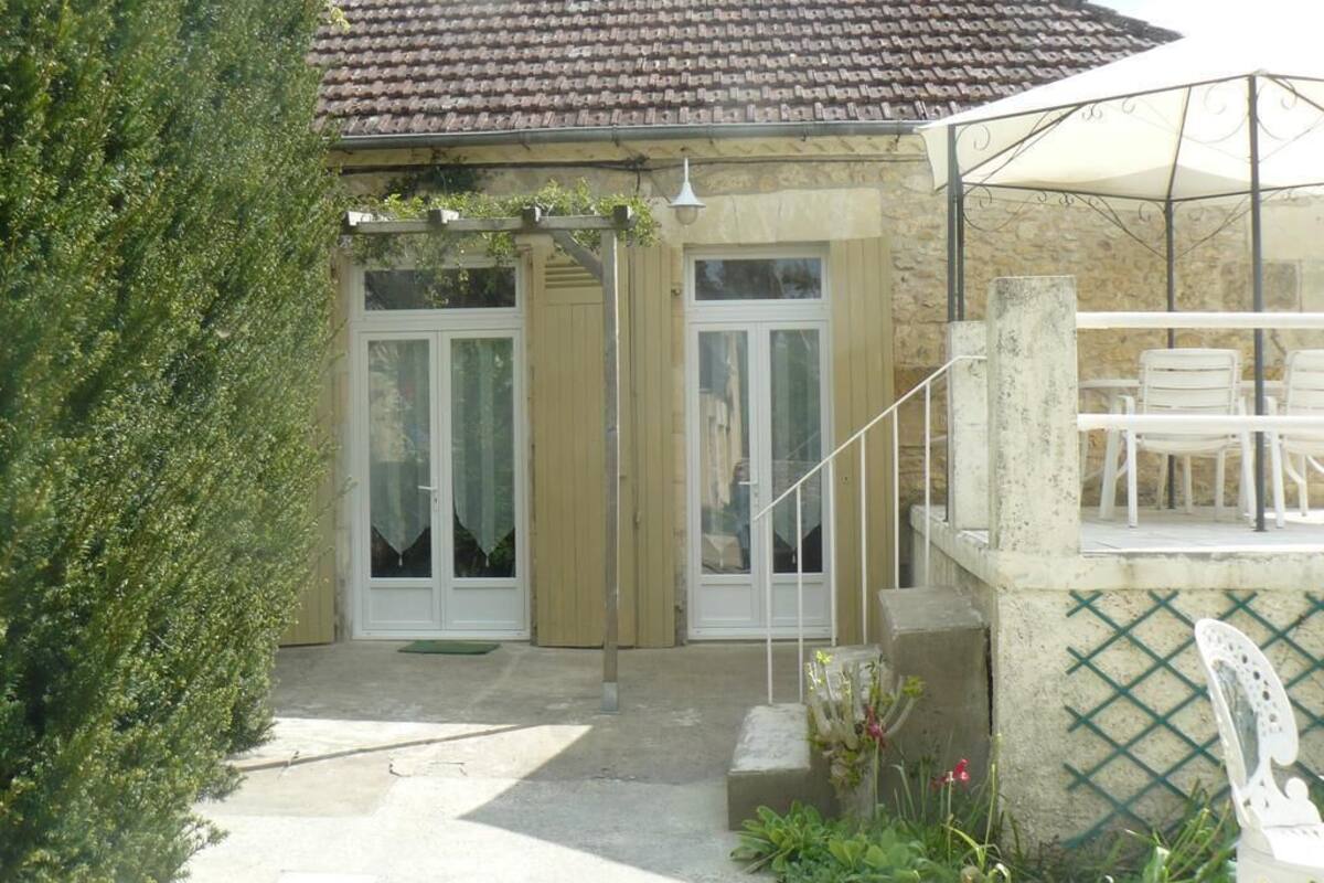 Un bâtiment doté d’un toit en tuiles, d’un patio couvert avec des chaises blanches et d’un petit jardin agrémenté de plantes et de fleurs.
