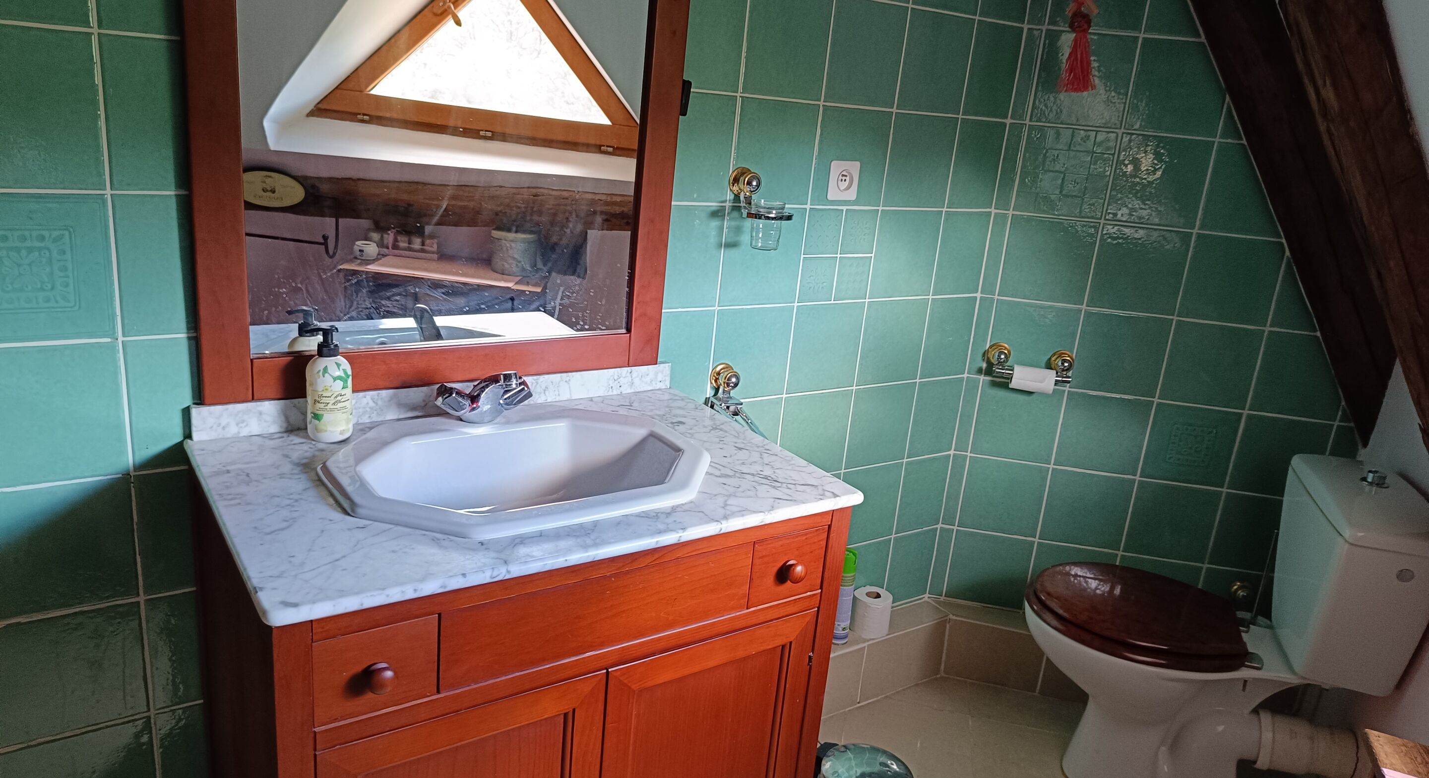 A bathroom with a sink, mirror, wooden cabinet, toilet, and green tiled walls.