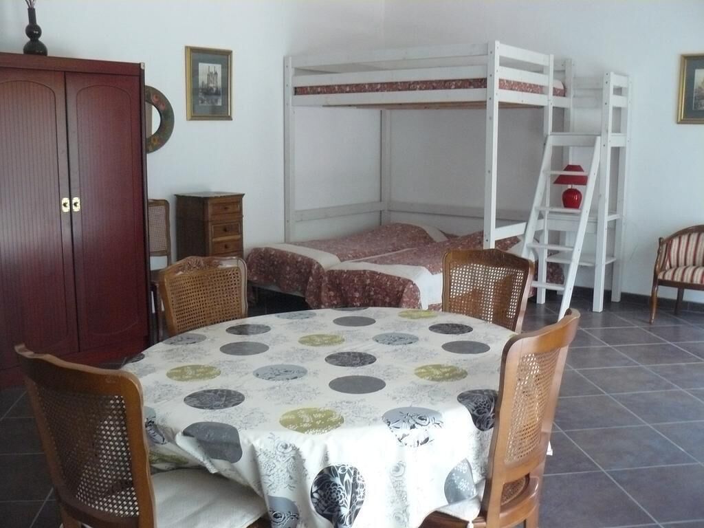 A room with a bunk bed, a dining table with a patterned tablecloth, wooden chairs, a cabinet, and a ladder.